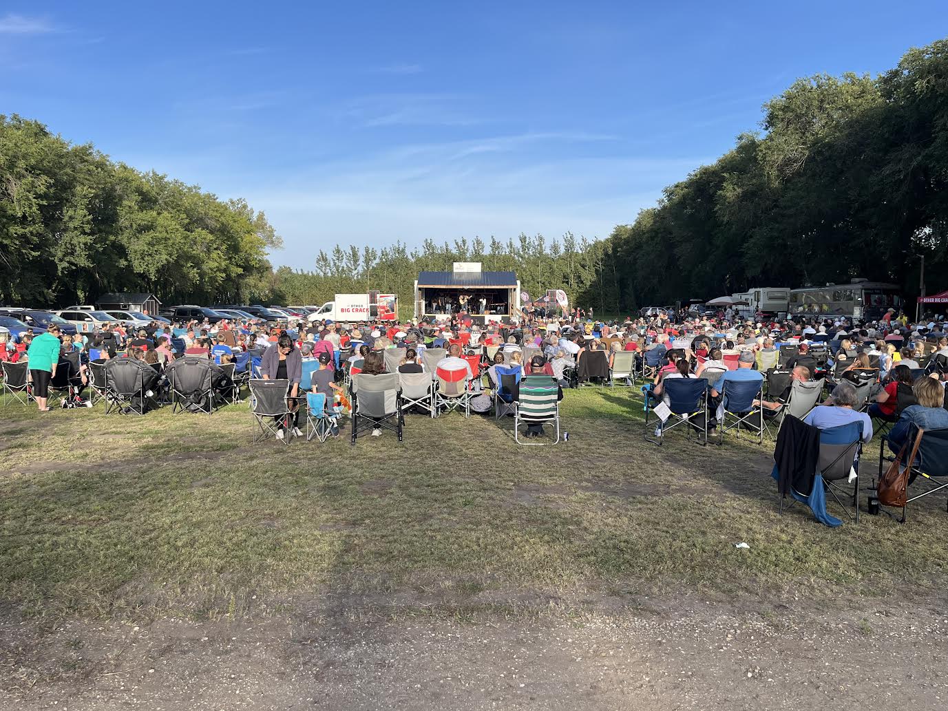 Lawn chairs set up in a field with trees on either side. The sky is blue with a few clouds. Music stage in the background....