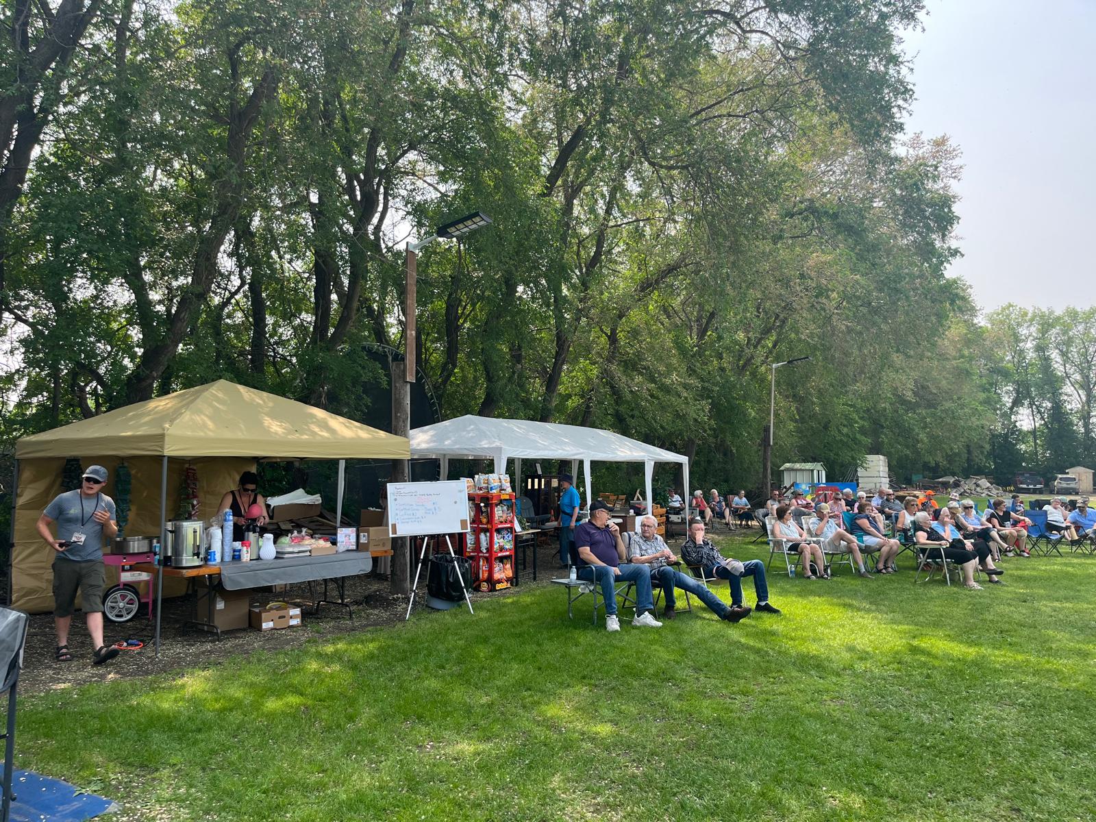 A group of people sit in lounge chairs on the grass, facing a gazebo with a whiteboard and counter, surrounded by trees in...