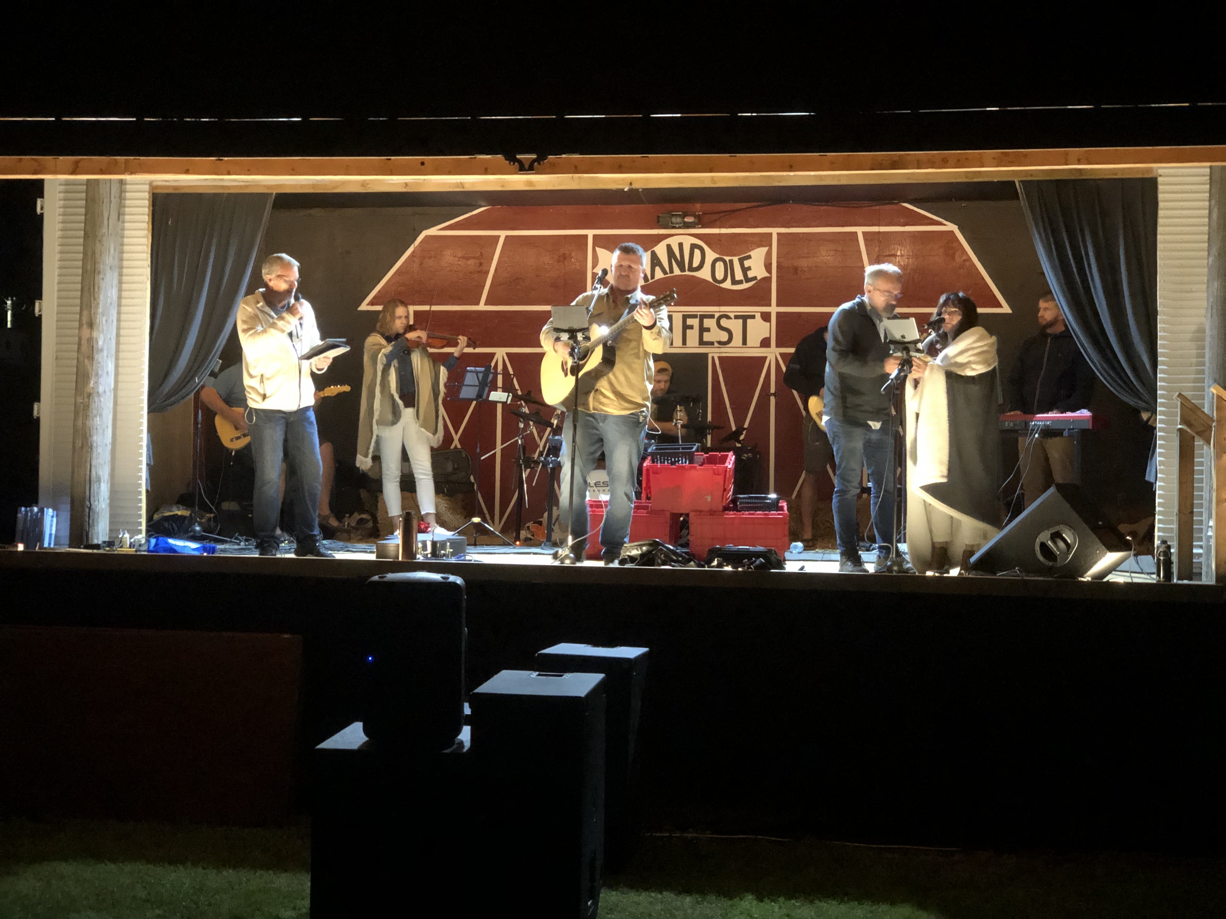 A group of performers are playing music on a stage with a red barn mural in the background that reads, "Grand Ole 'Ole FEST."